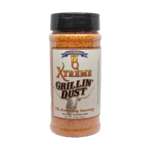 Bottle of B Xtreme BBQ Grillin Dust beside grilled steak, ribs, and corn on a wooden board