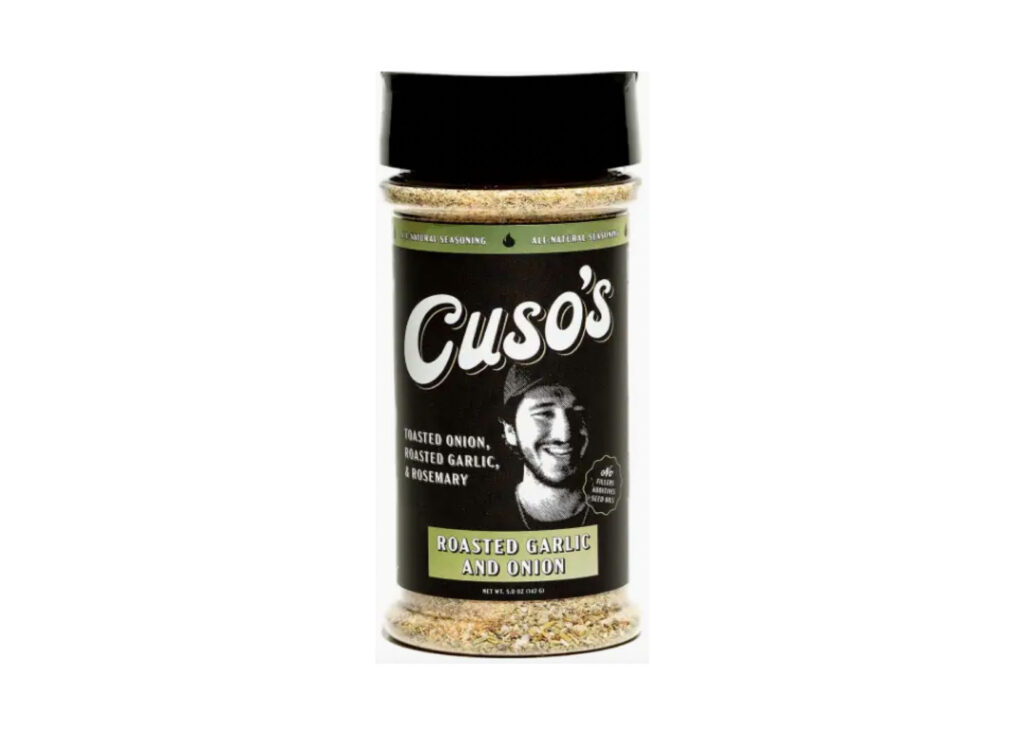 Bottle of Cuso’s Roasted Garlic & Onion Seasoning beside grilled steak and roasted garlic cloves on a wooden board